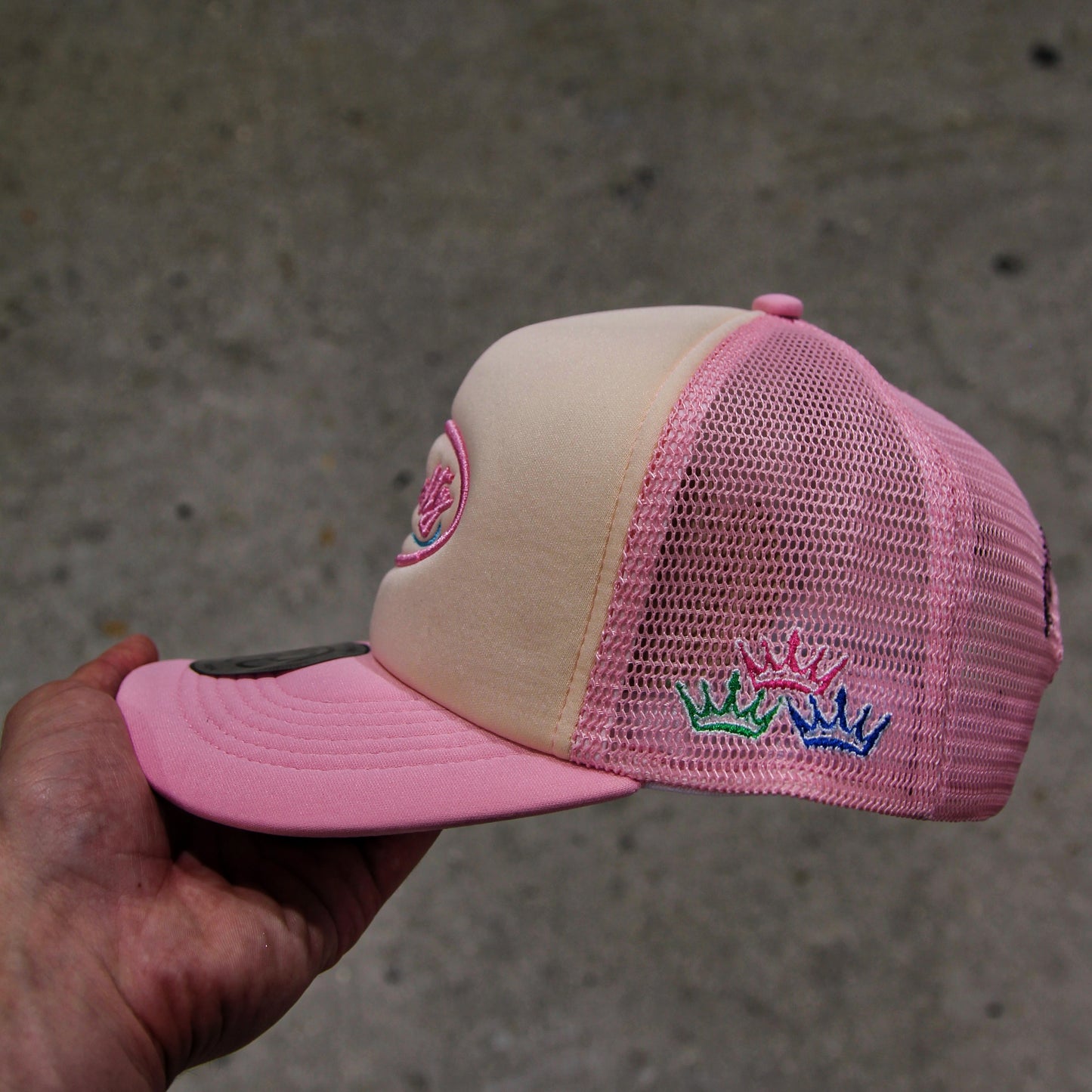 Pink and beige cap with embroidered design held by a hand against a concrete background