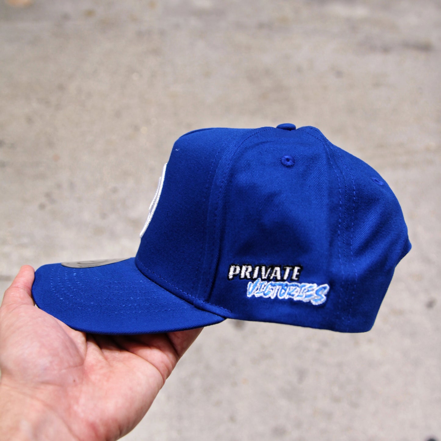 Blue cap with 'PRIVATE VICTORIES' text held by a hand on a concrete surface