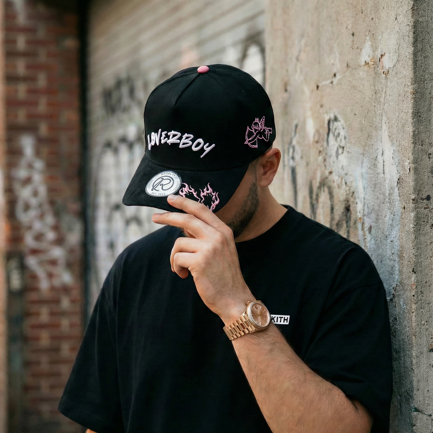 Man wearing a black cap with text and graphics, leaning against a wall with graffiti.