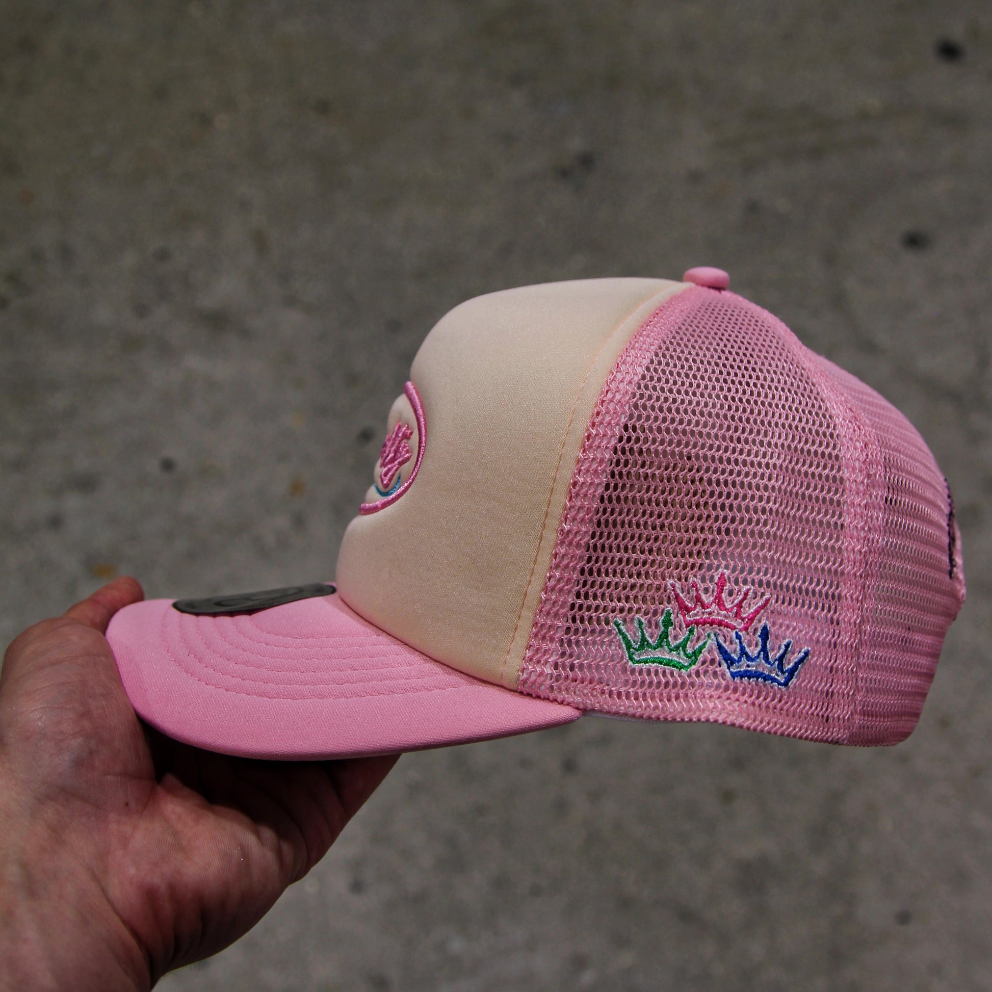 Pink and beige cap with embroidered design held by a hand against a concrete background