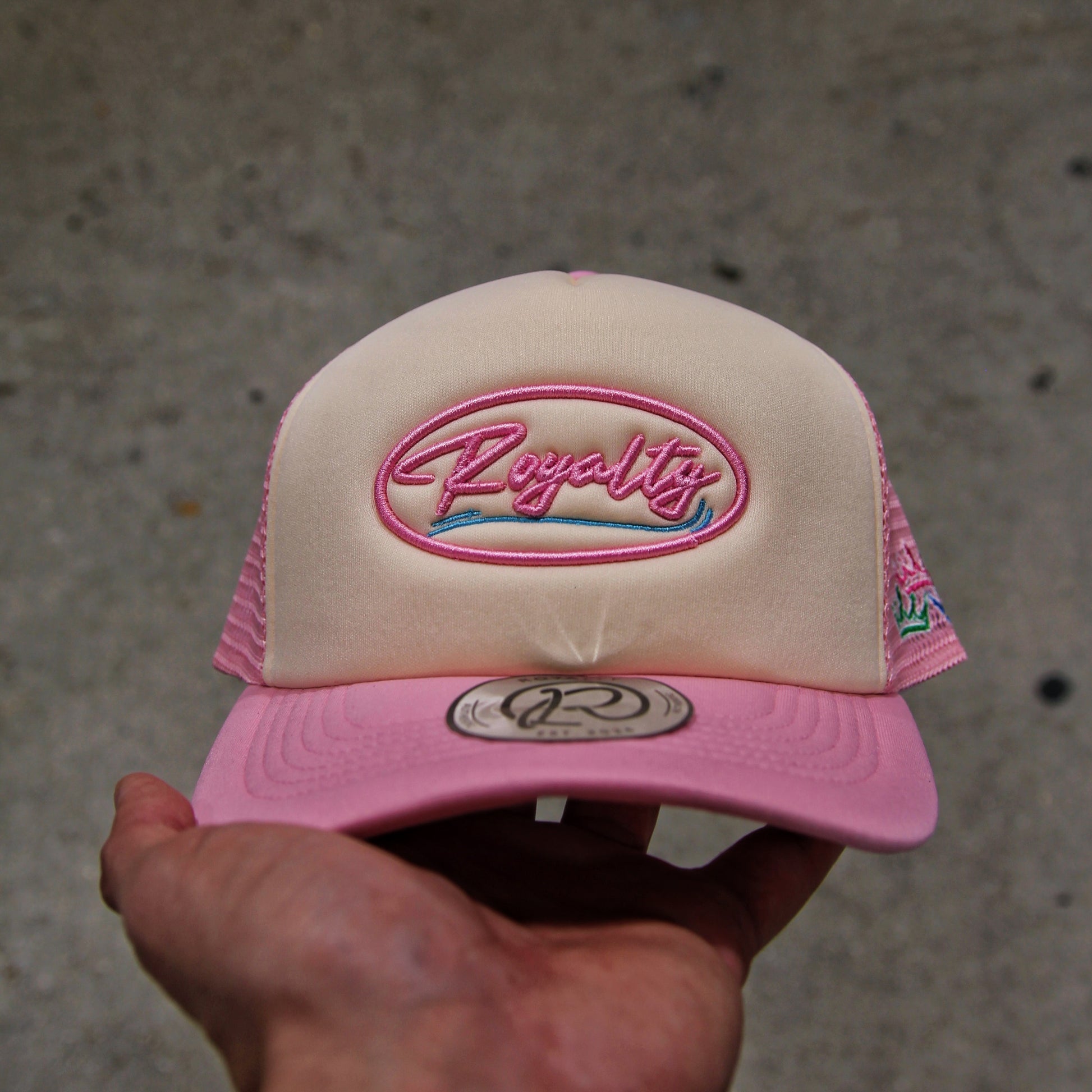 Pink and beige cap with 'Royalty' logo held by a hand on a concrete surface