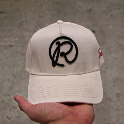 Beige cap with black logo held by a hand against a concrete wall background